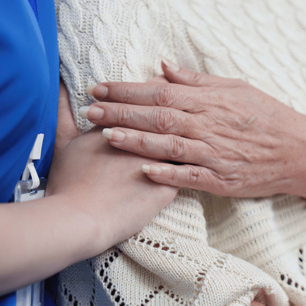 A close-up of an elderly hand resting gently on top of a younger person's hand, both on a cream-colored knitted blanket, symbolizing care, comfort, and support.