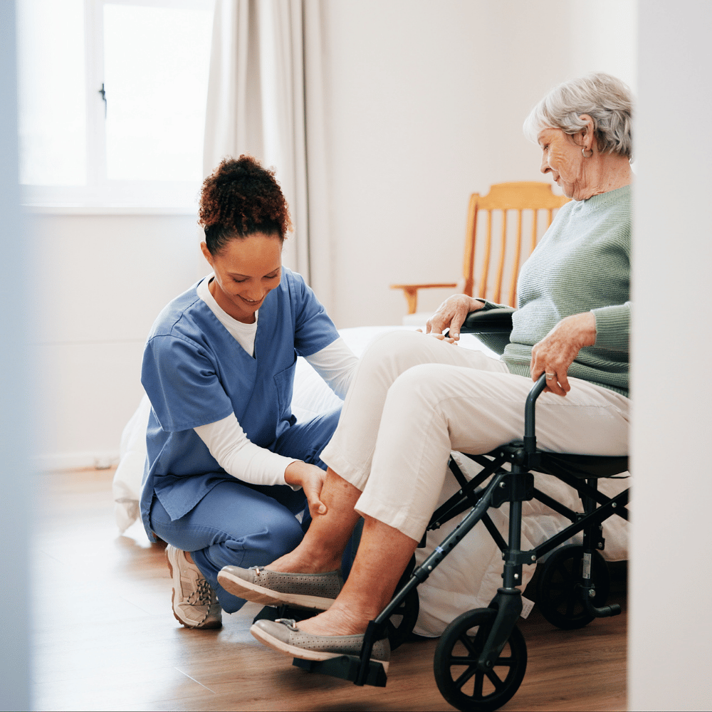 A nurse in blue scrubs helps an older woman in a green sweater, who is sitting on a wheelchair near a bed in a bright room. The nurse is adjusting the woman's leg rest and both appear calm and focused.