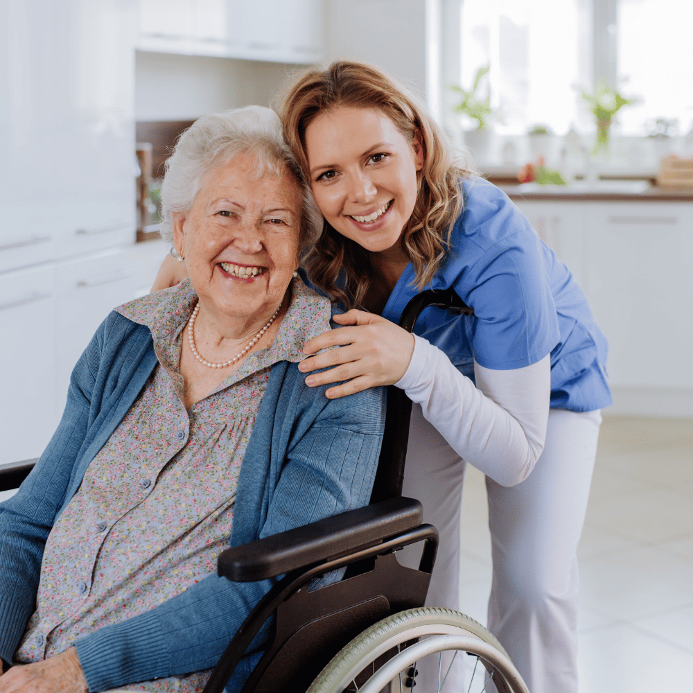An elderly woman in a wheelchair smiles while a young caregiver in blue scrubs stands beside her, also smiling, in a bright, modern kitchen.